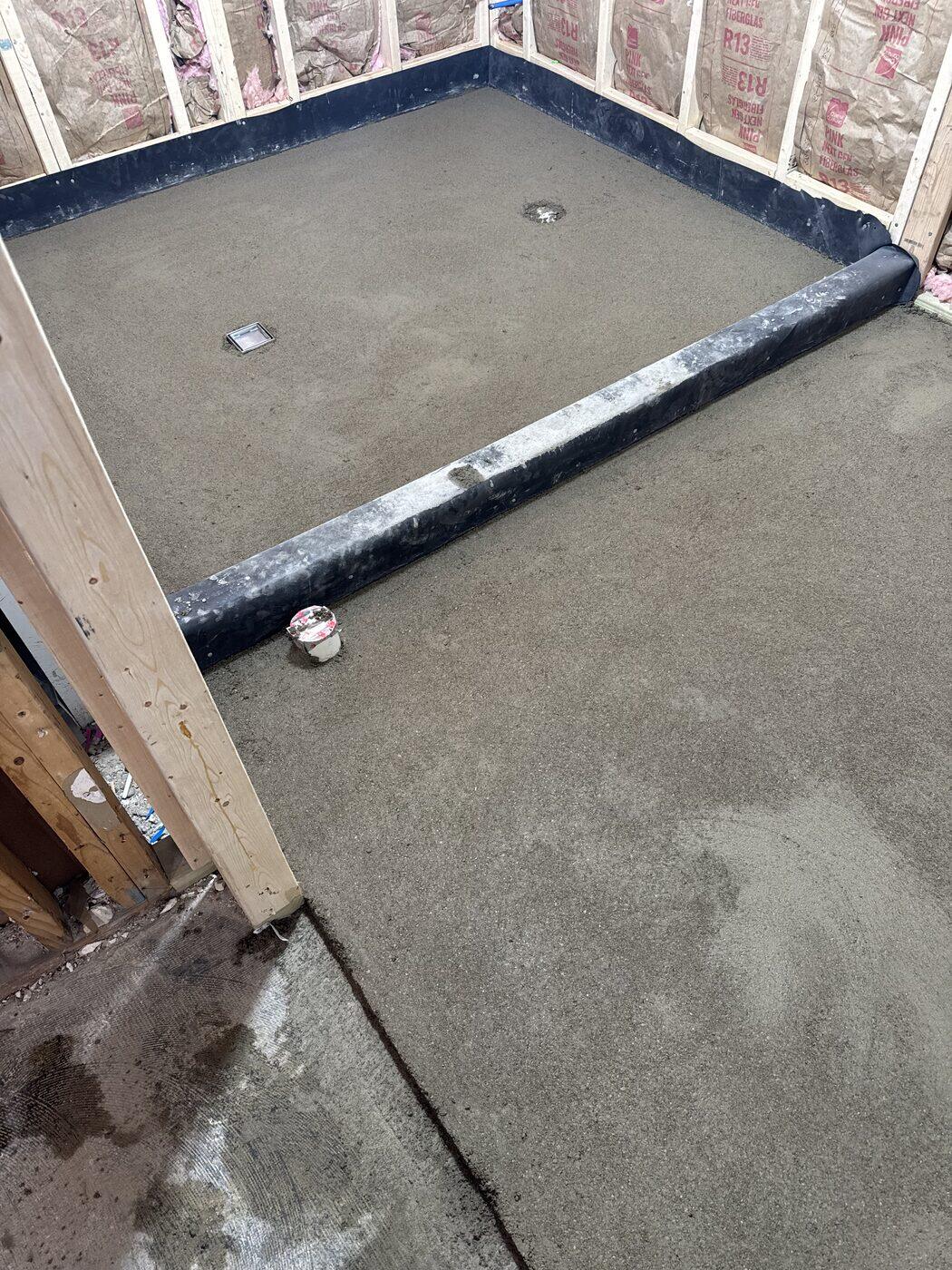 A wet-room floor floated over black waterproofing with a wrapped curb and two flush drains.