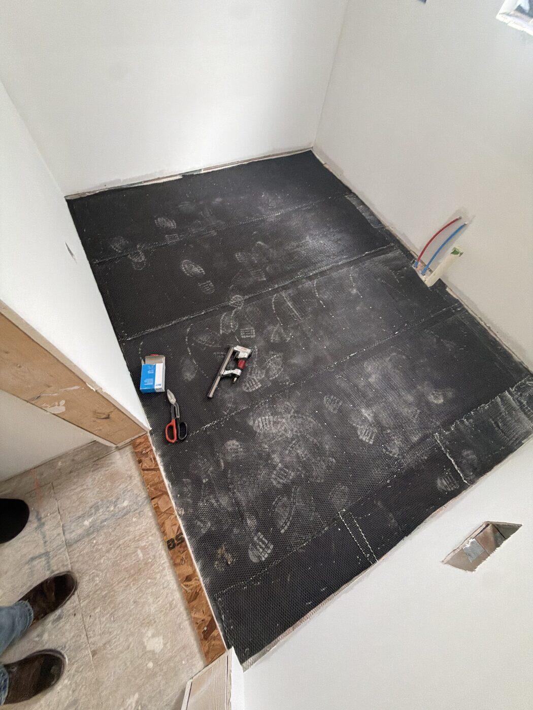 Black uncoupling membrane laid and rolled flat over a floor with cutting tools resting on it.